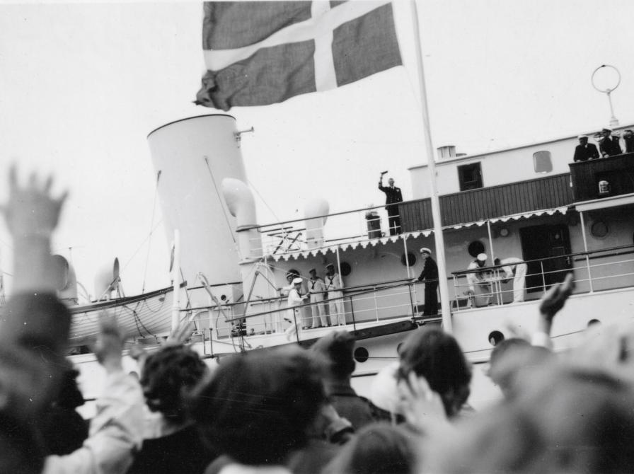 Sort-hvid foto af kongeskibet med Kong Frederik, der vinker fra dækket. Menneskemængde på kajen.